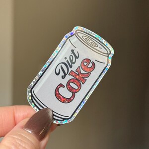May include: A Diet Coke sticker in the shape of a soda can. The can is white with the words "Diet Coke" in a cursive font. The text "Coke" is in red glitter. The sticker has a holographic border.