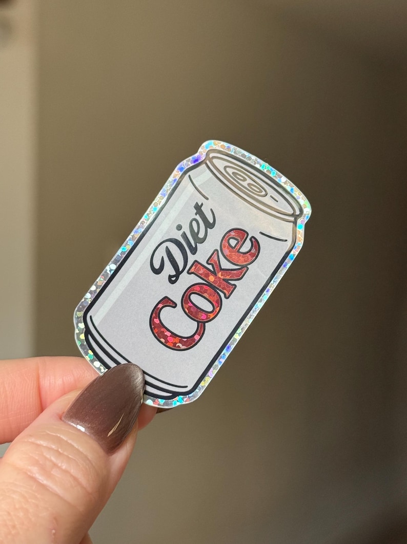 May include: A Diet Coke sticker in the shape of a soda can. The can is white with the words "Diet Coke" in a cursive font. The text "Coke" is in red glitter. The sticker has a holographic border.