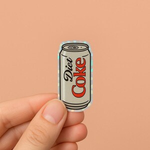 May include: A Diet Coke sticker in a can shape. The can is silver with the words "Diet Coke" in black and red. The sticker has a holographic, iridescent finish.