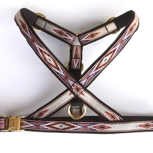 May include: A black dog harness with a white, red, and blue geometric pattern. The harness has a metal buckle and D-ring.