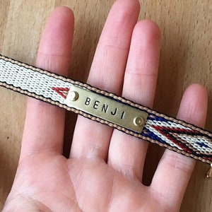 Può includere: Un braccialetto intrecciato con una placca in metallo dorato incisa con il nome "BENJI". Il braccialetto ha un motivo intrecciato in nero, bianco, rosso e blu.