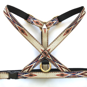 Op de afbeelding: Een beige hondenharnas met een geometrisch patroon in tinten rood, blauw en zwart. Het harnas heeft een zwarte verstelbare band en een gouden metalen ring.