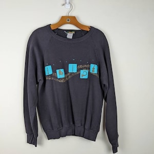 Peut inclure: Un sweat-shirt noir avec un motif d'instruments de musique en bleu et or sur le devant. Le motif dit "April Music".