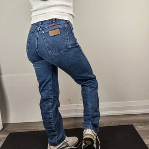 May include: A person wearing a white t-shirt and blue denim jeans with a brown leather patch on the back pocket. The person is wearing white sneakers with yellow accents.