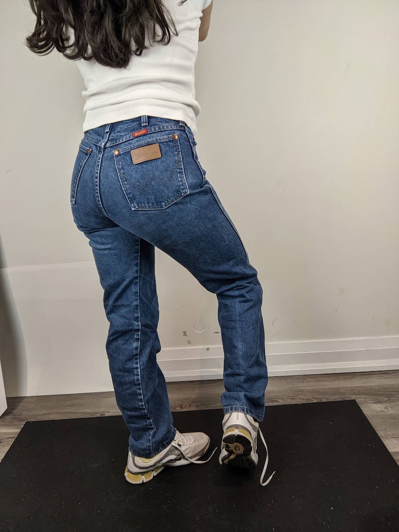 May include: A person wearing a white t-shirt and blue denim jeans with a brown leather patch on the back pocket. The person is wearing white sneakers with yellow accents.