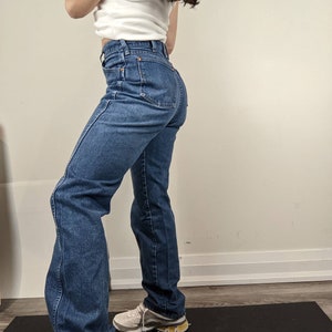 May include: A pair of blue denim jeans with a high waist and a straight leg fit. The jeans are worn by a person standing on a black yoga mat.
