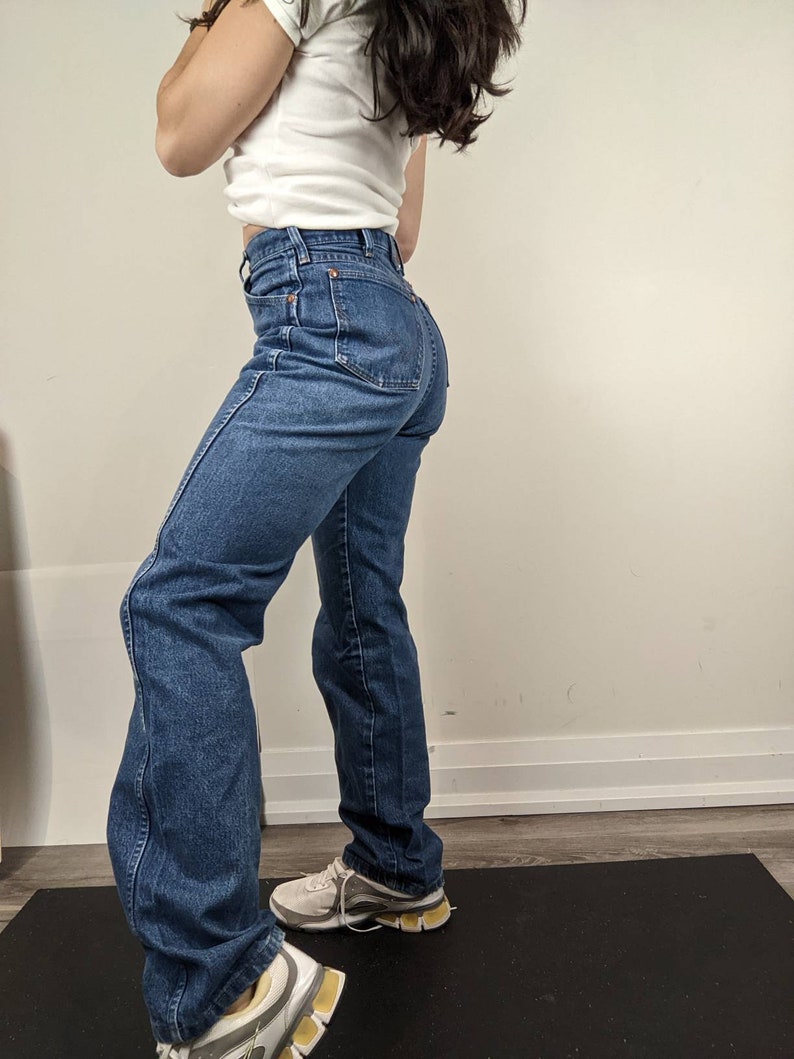 May include: A pair of blue denim jeans with a high waist and a straight leg fit. The jeans are worn by a person standing on a black yoga mat.