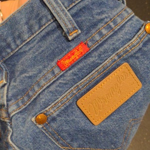 May include: A pair of blue denim jeans with a brown leather patch that says "Wrangler" and a red patch that says "Wrangler" on the back pocket.