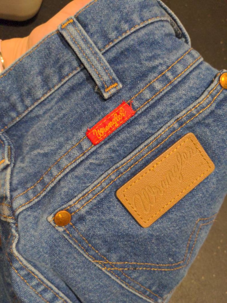 May include: A pair of blue denim jeans with a brown leather patch that says "Wrangler" and a red patch that says "Wrangler" on the back pocket.