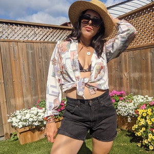 Könnte beinhalten: Eine Frau trägt ein weiß-braunes gemustertes Hemd, das in der Taille gebunden ist, schwarze Jeans-Shorts, einen Strohhut und eine Sonnenbrille. Sie steht in einem Garten mit Blumen.