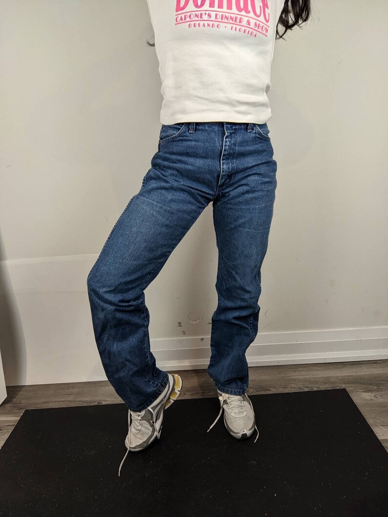 May include: A person wearing a white t-shirt with the text "Capone's Dinner & Show Orlando, Florida" in pink lettering, blue jeans, and gray sneakers.