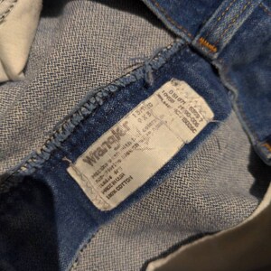 May include: A blue denim Wrangler jeans label with white text. The label says "Wrangler", "Made in USA", "100% Cotton", "13/10", "9X3", "10458 2190-006", "051071: 55097", "150260", "Machine wash with cold water", "Non-chlorine bleach", "Tumble dry low", "Do not iron".