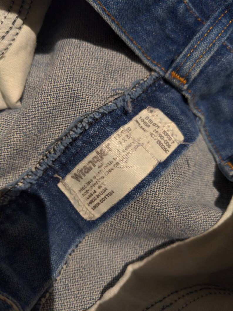 May include: A blue denim Wrangler jeans label with white text. The label says "Wrangler", "Made in USA", "100% Cotton", "13/10", "9X3", "10458 2190-006", "051071: 55097", "150260", "Machine wash with cold water", "Non-chlorine bleach", "Tumble dry low", "Do not iron".
