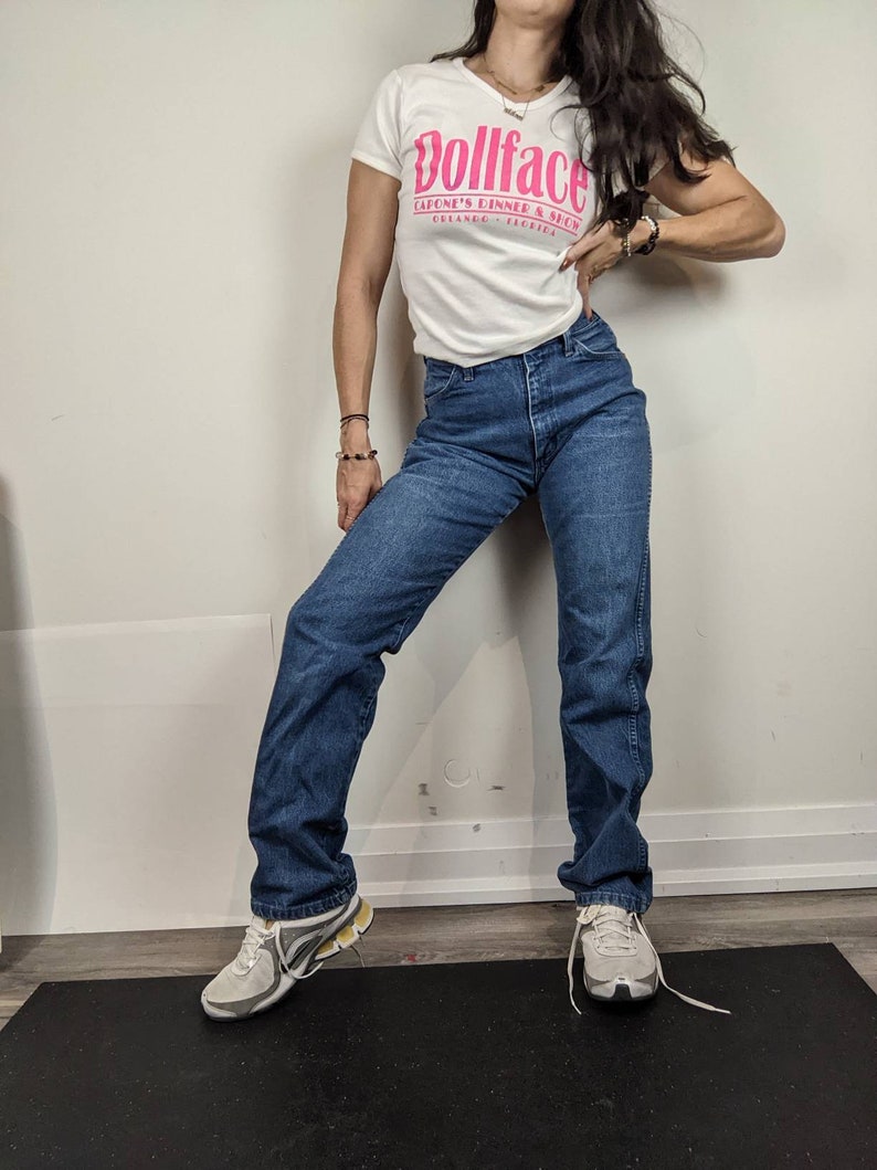 May include: A woman wearing a white t-shirt with a pink graphic that says "Dollface Capone's Dinner & Show Orlando, Florida" and blue jeans. She is wearing white sneakers.