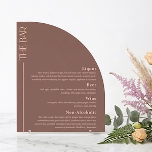 May include: A brown semi-circular bar menu with a list of liquor, beer, wine, and non-alcoholic beverages. The text "THE BAR" is at the top left. The menu is propped up on a clear stand, with flowers in the background.