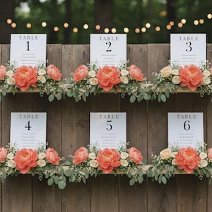 May include: Six white table number signs with black text, each adorned with floral arrangements of coral peonies, cream roses, and greenery. The signs are mounted on a wooden backdrop, creating a rustic and elegant display for a wedding or event.