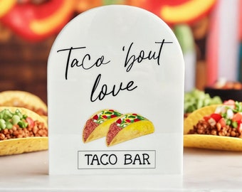 May include: White sign with black text that reads "Taco 'bout love Taco Bar". The sign has a picture of two tacos on it.