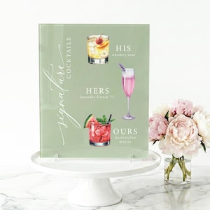 May include: A light green acrylic sign with the words "signature cocktails" and illustrations of three cocktails: a whiskey sour, a lavender french 75, and a watermelon mojito. The sign is on a white cake stand.