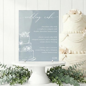 May include: A light blue sign with the words "wedding cake" in script. The sign lists cake flavors: chocolate with raspberry compote, carrot with walnuts, and vanilla with coconut cream. A white cake stand holds the sign, with greenery at the base.