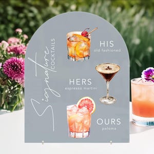 May include: A gray sign with the text "Signature Cocktails" in white script. The sign features illustrations of three cocktails: an old fashioned, an espresso martini, and a paloma. The text below each cocktail reads "His old fashioned", "Hers espresso martini", and "Ours paloma".