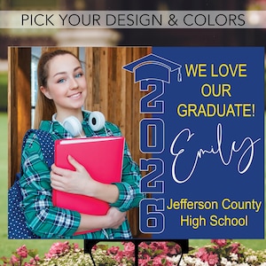 May include: A graduation yard sign with a blue background and the text "WE LOVE OUR GRADUATE!" and "2026". The sign features a photo of a smiling student holding a red folder. The sign also includes the name "Emily" and "Jefferson County High School".
