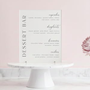 May include: A white dessert bar sign with a list of treats, including cupcakes, doughnuts, brownies, and cookies. The sign is on a white marble cake stand, with a pink background.