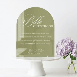 May include: An olive green acrylic Bible guestbook sign with an arched top. White script reads "Bible Guestbook" and invites guests to write notes. The names "Jackie & Rob" and the date "May 15, 2026" are also displayed. The sign is on a white cake stand.