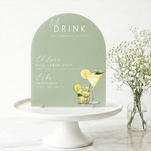 May include: A sage green arched sign with the text "let's DRINK" and "our signature cocktails". Below are the names and ingredients of two cocktails, "Chelseas" and "Erika". The sign is on a white cake stand, with a vase of white flowers.