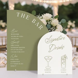 May include: Two decorative signs for a bar, one olive green and one white, with the words "THE BAR" and "Signature Drinks" in elegant script. The green sign lists beer, spirits, and wine options. The white sign features illustrations of an espresso martini and a classic mojito.