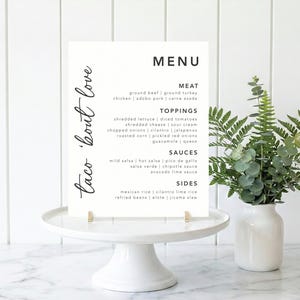 May include: A white menu card with the words "taco 'bout love" in cursive script. The menu lists meat, toppings, sauces, and sides, including ground beef, shredded lettuce, and mild salsa. The card is on a white cake stand.