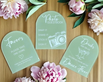 May include: Three light green acrylic signs with rounded tops and white text. The signs read "Photo Guestbook", "Favors Please Take One Home", and "Drinks Are On Us As Long As You're On The Dance Floor".