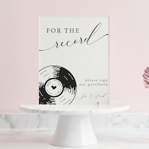 May include: A white sign with black text and a vinyl record graphic. The sign reads "FOR THE record" in script, with additional text below. The sign is on a white cake stand, ideal for a wedding or event.