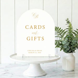 May include: An ivory-colored wedding sign with an arched top, displaying the words "CARDS and GIFTS" in gold lettering. The sign also includes the names "CHELSEA & BRIAN" and the date "MARCH 14, 2026". The sign is displayed on a white cake stand.