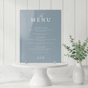 May include: A light blue acrylic menu with white text, displayed on a white cake stand. The menu lists appetizers, entrées, and desserts. A white vase with greenery is to the right.