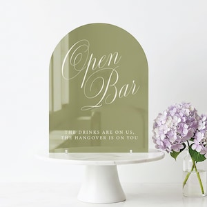 May include: An olive green acrylic sign with the words "Open Bar" in elegant white script. The sign has an arched top and is displayed on a white cake stand. The text "THE DRINKS ARE ON US, THE HANGOVER IS ON YOU" is at the bottom.