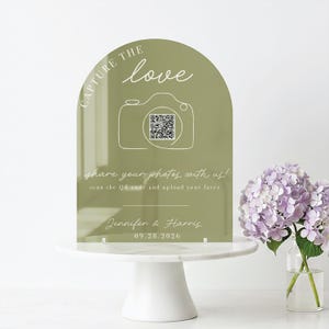 May include: An olive green acrylic wedding sign with the words "CAPTURE THE love" and a camera graphic. The sign includes a QR code and text inviting guests to share photos. It is displayed on a white cake stand with a bouquet of purple flowers.