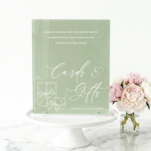 May include: Sage green acrylic sign with white script text that reads "Cards & Gifts." The sign includes the text "We are so grateful..." and the names "Heather & Graham." The sign is on a white cake stand.