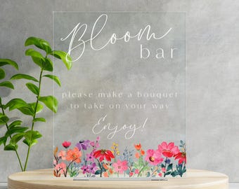 Könnte beinhalten: Ein klares Acrylschild mit dem Text "Bloom bar" und der Botschaft "Bitte nehmen Sie einen Strauß mit. Genießen Sie!" Das Schild ist mit einem Aquarell-Blumenrand aus rosa, roten und violetten Blumen verziert.