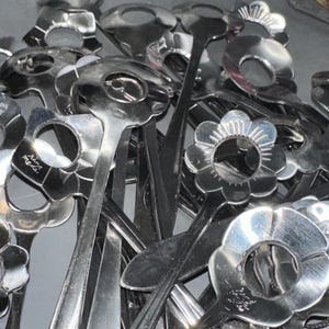 May include: A collection of shiny, silver-colored serving spoons. Each spoon features a flower-shaped head with a central opening and a long, slender handle. The spoons are clustered together, creating a reflective surface.