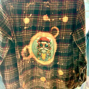 May include: A long-sleeved, button-down flannel shirt with a plaid pattern in shades of black, brown, and red. The back features a circular graphic of a bearded character with sunglasses. The shirt has a distressed, bleached effect.