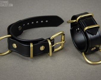 May include: A black leather collar and matching wrist cuffs with brass hardware. The collar has a D-ring and a buckle closure. The cuffs have a D-ring on one end and a buckle closure on the other.