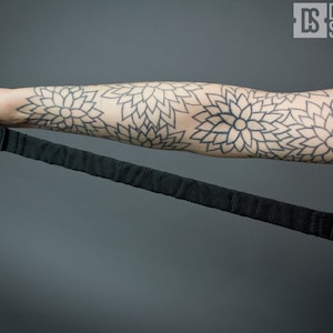 May include: A black adjustable strap with a padded handle. The strap is being held by a person with a black and white geometric tattoo on their arm.