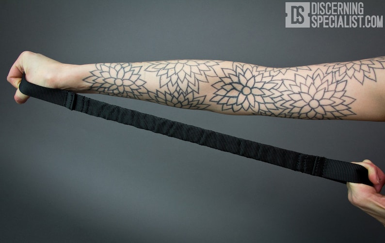May include: A black adjustable strap with a padded handle. The strap is being held by a person with a black and white geometric tattoo on their arm.