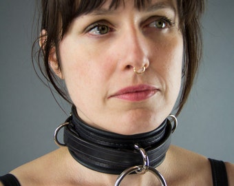 May include: A black leather choker with a silver ring. The choker is worn by a person with brown hair and a nose ring.