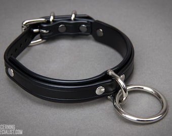 May include: Black leather collar with a silver metal ring and buckle. The collar has a rounded design with a central groove and is secured with rivets. The image includes the text "DISCERNING SPECIALIST.COM".
