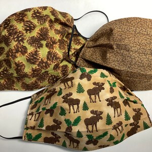 May include: Three fabric face masks with different woodland patterns. One mask features brown pine cones on a green background, another has a brown and tan speckled pattern, and the third has brown moose silhouettes on a tan background with green pine cones and ferns.