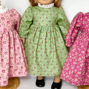 May include: Three doll dresses in pink, green, and fuchsia, each with a floral pattern. The dresses have long sleeves and are displayed on mannequins. Each dress has a white lace collar and a gathered skirt. The dresses are suitable for a doll.
