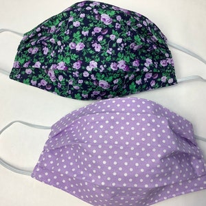 May include: Two fabric face masks. One mask is purple with white polka dots. The other mask is black with a floral pattern in shades of purple and green.