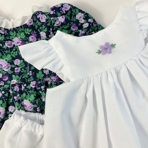 May include: Two baby dresses, one with a white bodice and ruffled sleeves, and the other with a dark blue bodice with a floral print and white ruffled sleeves.