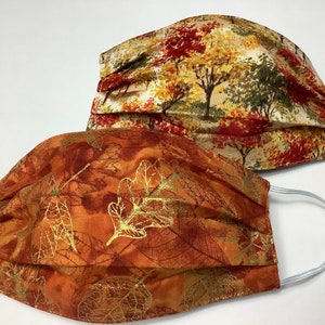 May include: Two fabric face masks with fall-themed prints. One mask features a brown, orange, and gold leaf pattern. The other mask features a white background with brown, orange, and green trees.